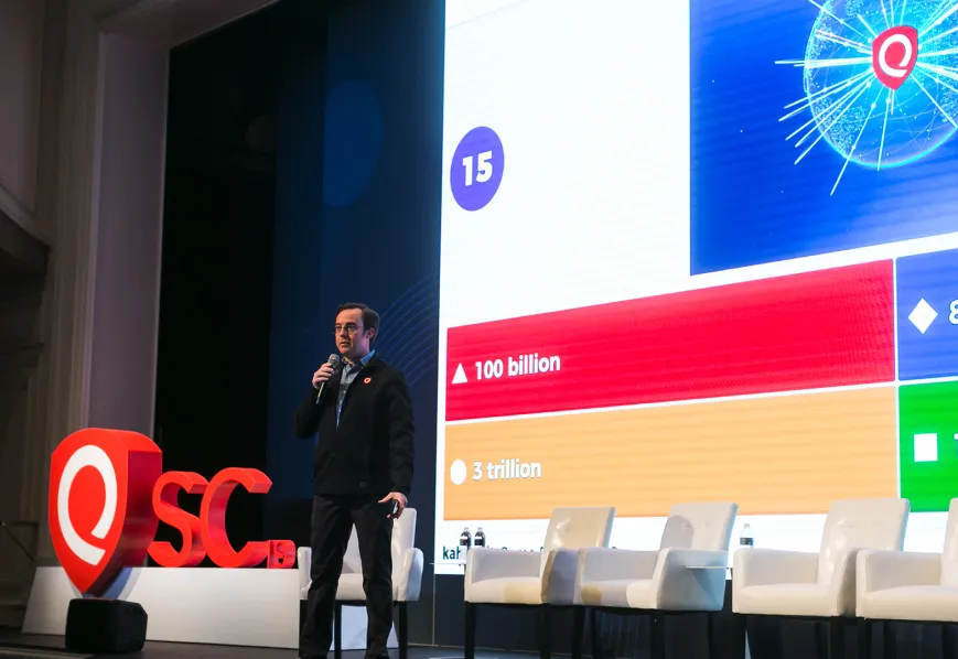 A speaker presenting on a stage next to a large screen displaying a live Kahoot! question. The stage features a row of white chairs and a large "QSC 19" logo.