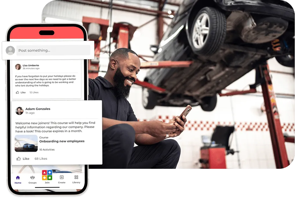 Smiling mechanic in a black uniform looking at his smartphone while crouching in a garage with a car lifted on a red hydraulic lift behind him.