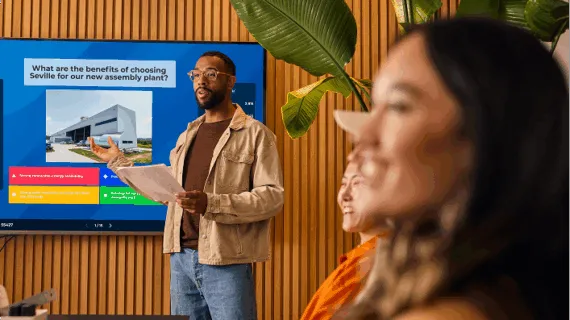 A male presenter leading an interactive business meeting, standing next to a large screen displaying a quiz question about the benefits of choosing Seville for a new assembly plant.