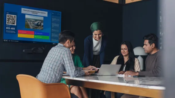 A diverse group of professionals collaborating around a laptop in a meeting room, with a large screen displaying a Kahoot! presentation in the background.