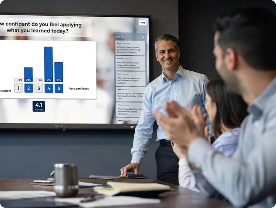A male presenter in a blue shirt smiles as a group of colleagues applauds in a boardroom. The large screen behind him shows a Kahoot! poll.