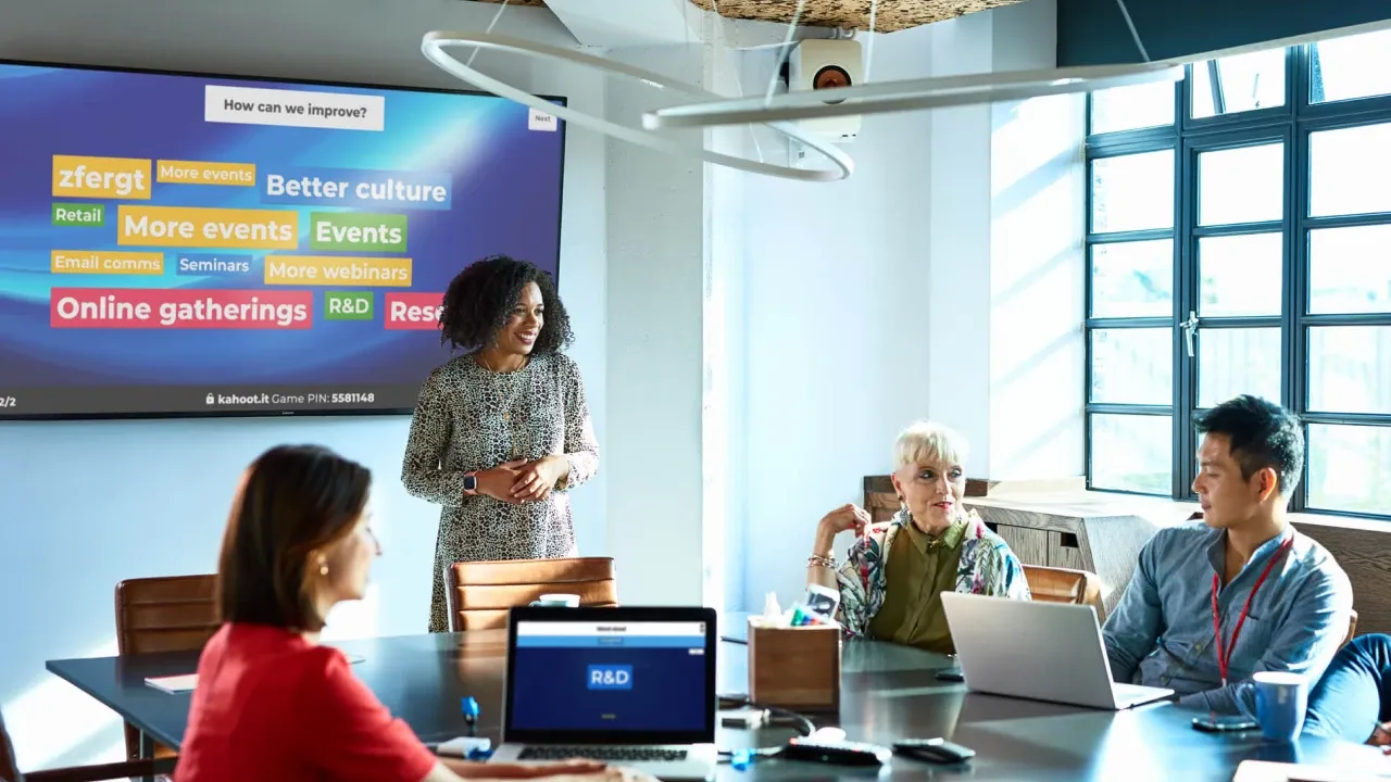 A presenter leads a team meeting using a Kahoot word cloud displayed on a large screen