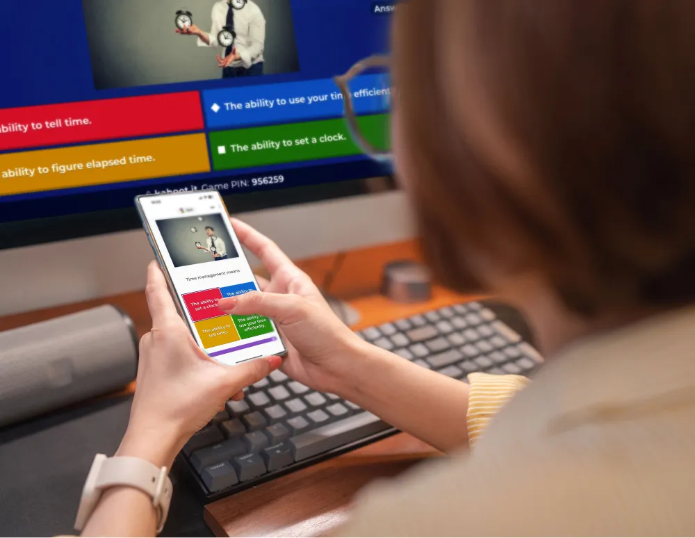 A person participates in an interactive kahoot quiz on their smartphone while sitting at a desk. The phone screen shows a question about time management with multiple-choice answers, and a larger screen in the background displays the same quiz.