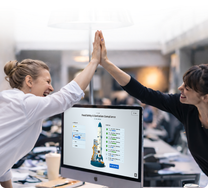 Two female colleagues in a modern office smile and share a high-five over a desktop monitor.
